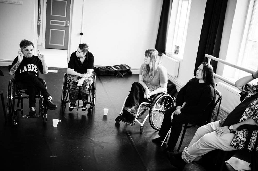 Svartvit bild av en samtalscirkel i ett ljust rum. Flera deltagare sitter på stolar, några i rullstol. En person gestikulerar medan de andra lyssnar. Kaffemuggar står på golvet i mitten av cirkeln.