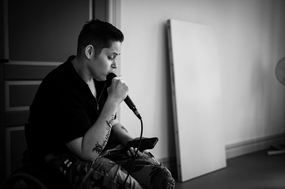 Svartvit närbild av en person som sitter och talar i en handhållen mikrofon medan hen läser från en mobiltelefon. Personen är koncentrerad och sitter i ett ljust rum med tom vägg i bakgrunden.