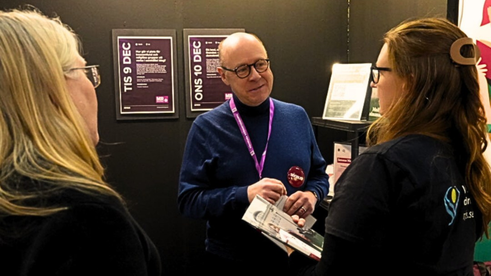 Tre personer samtalar vid en monter under MR-dagarna. En man med namnbricka håller i informationsmaterial och talar med två kvinnor. På väggen bakom syns affischer med programdatum i december.