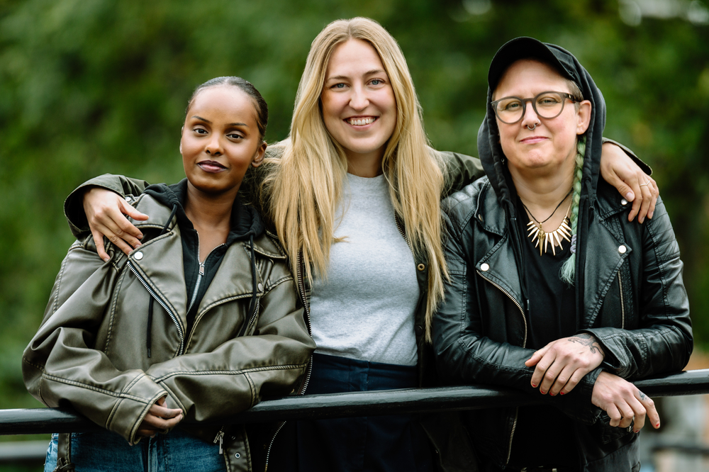 Tre personer står utomhus framför en suddig grön bakgrund. De står tätt intill varandra och ler mot kameran, med armarna lagda om varandras axlar.
