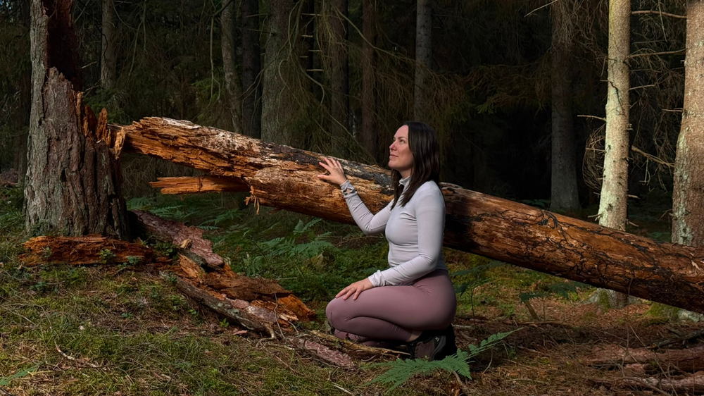 En kvinna sitter på huk i en mörk skog och rör vid ett fallen, vittrat trädstamme med ena handen. Hon blickar uppåt med ett lugnt uttryck, omgiven av mossbevuxen mark och granar.