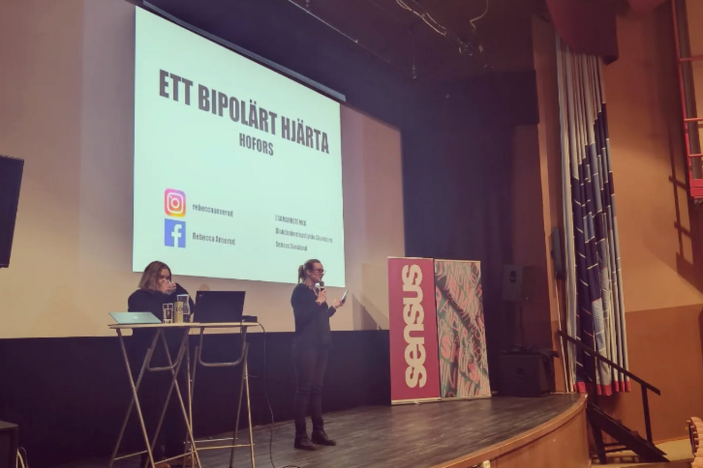 En person står på en scen och håller en presentation framför en stor projicerad bild med texten ”Ett bipolärt hjärta”. En annan person sitter vid ett bord med laptop. På scenen står även en roll-up med Sensus‑logotypen.