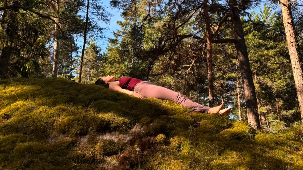 En kvinna ligger utsträckt på rygg på en solbelyst mossbevuxen kulle i skogen. Barfota och med armarna längs kroppen utstrålar hon djup avslappning, omgiven av tallar och blå himmel.