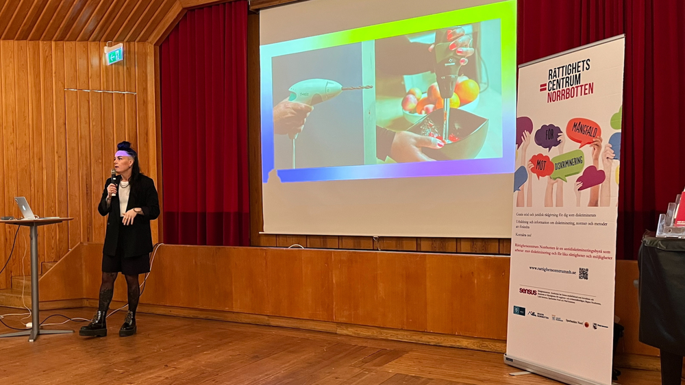 Mariana Vnuk föreläser på scen med mikrofon under ett arrangemang av Rättighetscentrum Norrbotten. Bakom henne visas en presentation på stor duk.