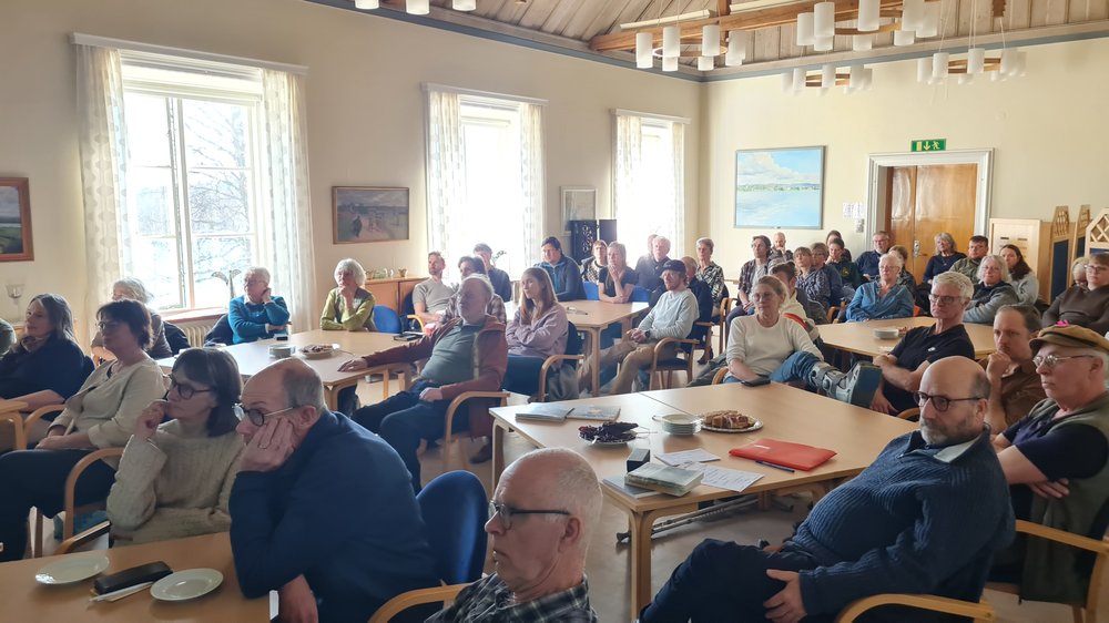 Ett trettiotal personer sitter samlade i en ljus lokal och lyssnar vid bord med kaffe och fika under ett möte eller samtal.