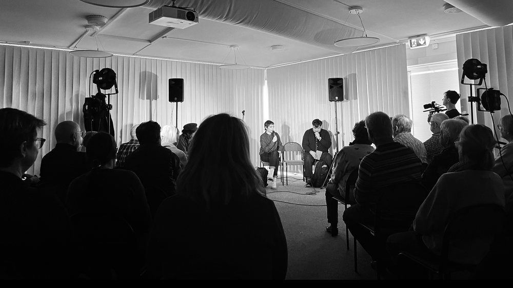 Svartvit bild av ett publikt samtal i en mindre sal. Två personer sitter längst fram med mikrofoner och talar inför publik. Högtalare, scenbelysning och en kameraman syns i rummet.   Ask  Explain