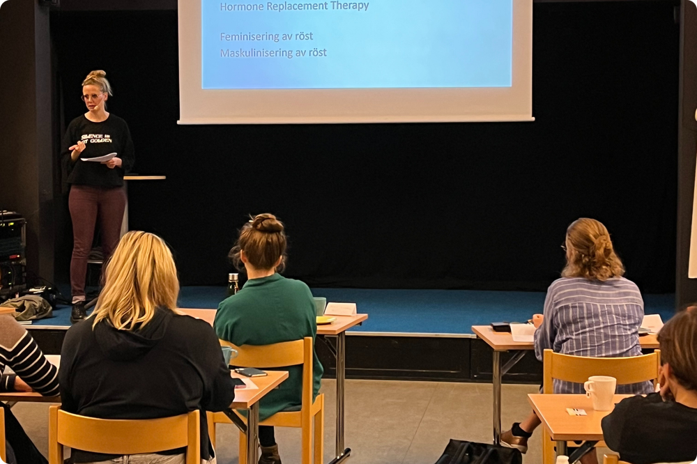 En kvinna föreläser för en grupp deltagare på en scen. Framför henne ett podie med en laptop och i bakgrunden i projektorduk. Framför scenen sitter kursdeltagare på bord och stolar.