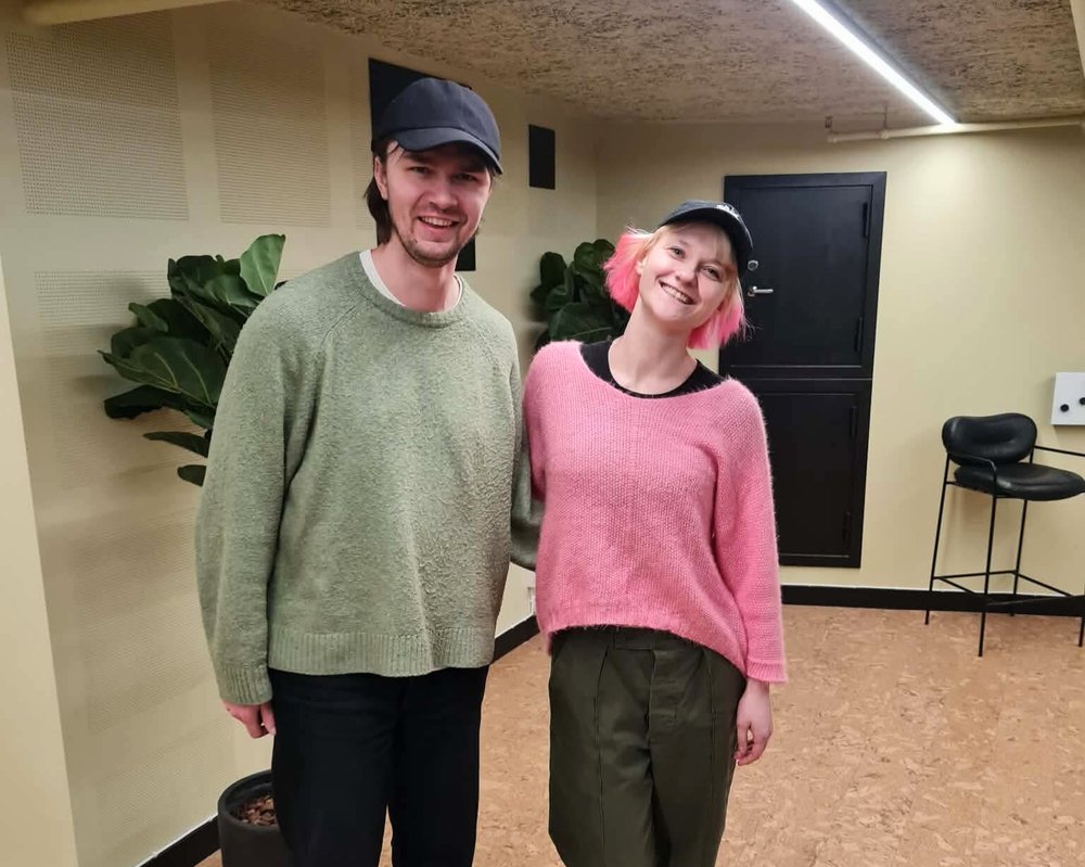 Två personer står i ett mötesrum och ler mot kameran under en låtskrivarcirkel. De deltar i gemensamt låtskrivande. Växter och stolar syns i bakgrunden.