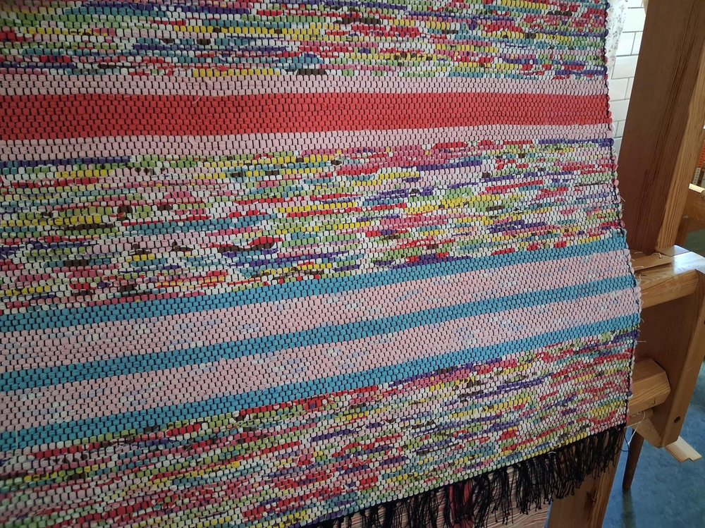 Närbild på en färgglad trasmatta i vävstol, med ränder i rosa, blått, rött och flerfärgade mönster.