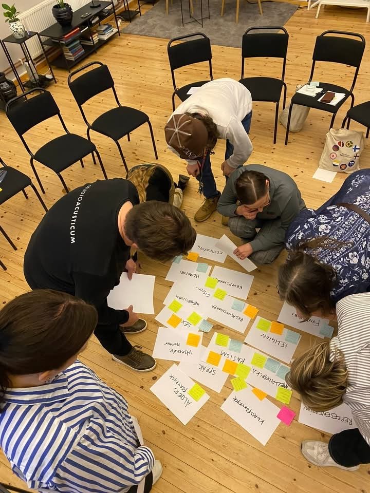Deltagare i en workshop sitter och står runt papper på golvet med ord och post-it-lappar. Gruppen arbetar tillsammans i en övning om normer, bemötande och att säga ifrån.